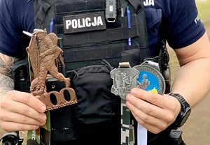 tułów policjanta w założonej kamizelce taktycznej z opisem policja, policjant trzyma statuetki otrzymane jako nagrody za udział w biegach