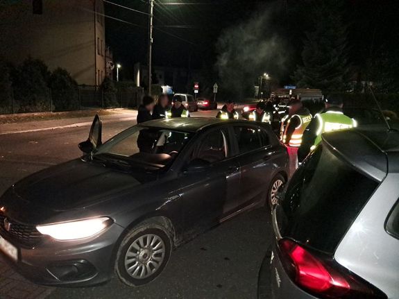 Zdjęcie w kolorze przedstawia w porze nocnej na pierwszym planie dwa pojazdy za którymi stoją funkcjonariusze policji w kamizelkach odblaskowych.