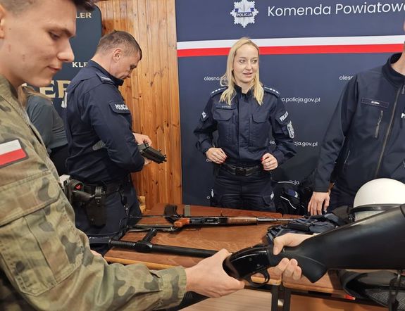 Zdjęcie w kolorze przedstawia stoisko policyjne z funkcjonariuszami, którzy promują służbę demonstrując uczniom jednostki broni i elementy wyposażenia.