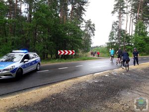 rowerzyści jadą trasą leśną, przejeżdżają przez jezdnię, po lewej stronie stoi  radiowóz po prawej stronie policjant