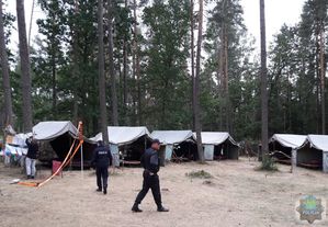 pole namiotowe, policjant i strażak chodzą po suchej ściółce, z lewej strony kobieta przyczepia kartkę do  tablicy informacyjnej, w tle szare namioty i drzewa.