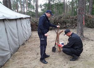 strażak  sprawdza legalizację gaśnicy, obok stoi policjant trzyma w dłoni szpadel, obok z lewej strony szary namiot w tle ogrodzenie z gałęzi iglastych w oddali drzewa.