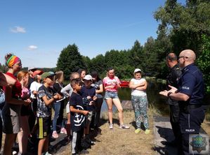 Policjant i strażak rozmawiają z dziećmi, świeci słońce, w tle zbiornik wodny po którym pływają kajaki.