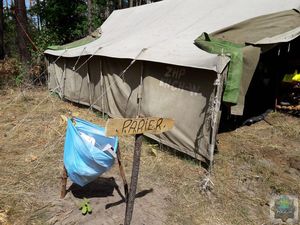 drewniana deska z napisem papier obok  plastikowy niebieski worek z papierem, na drugim planie szary namiot harcerski w tle las.