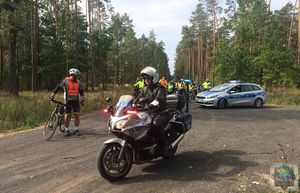 Uczestnicy rajdu rowerowego stoją na leśnej drodze przed nimi radiowóz i policjant na motocyklu.