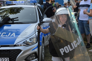 dziecko ubrane w sprzęt policyjny, tarcza, kamizelka, hełm, pałka. dziecko stoi obok radiowozu