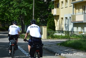 2 policjantów na rowerach jedzie ulicą obok blok mieszkalny