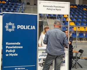 z lewej  strony  niebieskie tło  z napisem  Policja i  Komenda Powiatowa Policji  w Nysie, obok tła stoją trzy  osoby, pierwsza kobieta ubrana na biało, dalej stojący  tyłem  mężczyzna, ostatnia stoi  policjantka