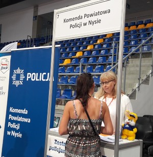 z lewej strony niebieski baner z napisem Policja i  Komenda Powiatowa Policji w Nysie, obok baneru  stoją dwie kobiety, pierwsza stoi  tyłem  w kolorowej  bluzce, druga ubrana na biało stoi przodem.