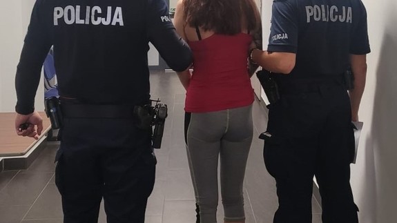 dwóch  policjantów prowadzi kobietę w czerwonej  bluzce.  Wszystkie osoby stoją tyłem