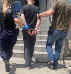 kolorowe zdjęcie - policjanci prowadza osobę zatrzymaną. Zatrzymany mężczyzna ma założone kajdanki na ręce.