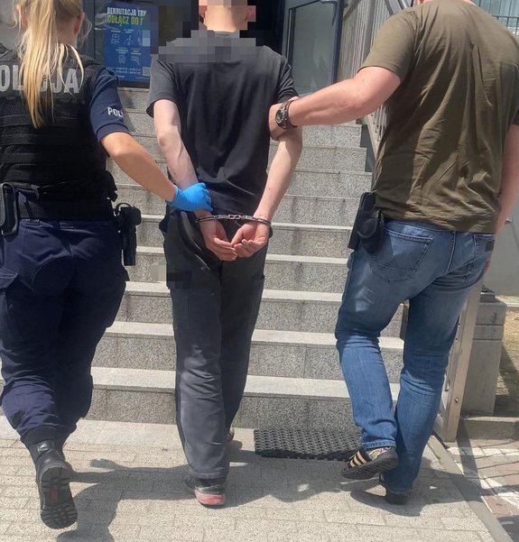 kolorowe zdjęcie - policjanci prowadza osobę zatrzymaną. Zatrzymany mężczyzna ma założone kajdanki na ręce.