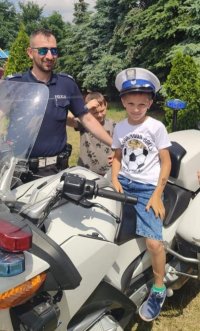 Policjant stoi przy motocyklu służbowym na którym siedzi chłopiec w policyjnej czapce