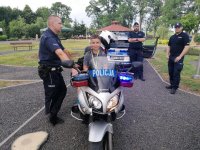 Policjant stoi obok służbowego motocykla na którym siedzi chłopiec, w tle widać dwóch innych policjantów i radiowóz