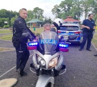 Policjant stoi obok służbowego motocykla na którym siedzi dziewczynka, w tle widać innego policjanta i radiowóz