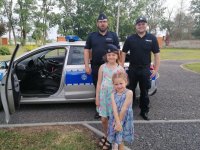 Przed radiowozem stoi dwóch policjantów wraz z dwoma dziewczynkami