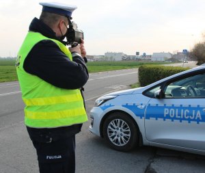 Umundurowany policjant na drodze kontroluje prędkość za pomocą ręcznego miernika pomiaru prędkości w tle widoczny oznakowany radiowóz.