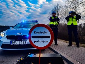 umundurowani policjanci w trakcie służby, kontrolują ruch na drodze. Obok radiowóz i znak z informacją o kontroli policyjnej.