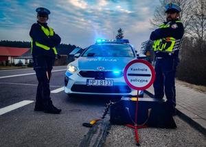 Umundurowani policjanci w trakcie służby, kontrolują ruch na drodze. Obok radiowóz i znak z informacją o kontroli policyjnej.