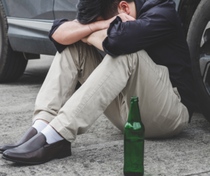 mężczyzna siedzi na jedni, opiera się o samochód, obok stoi zielona butelka