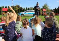 Na przedszkolnym podwórku policjantka rozmawia z dziećmi o zasadach bezpieczeństwa, w tle widać radiowóz