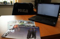 Na polarze napis policja, na biurku stoi laptop i leżą materiały promujące dobór do służby w Policji
