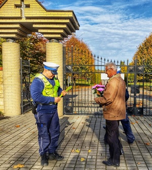 Zdjęcie przedstawia policjantów rozdając odblaski pieszym przed cmentarzem.