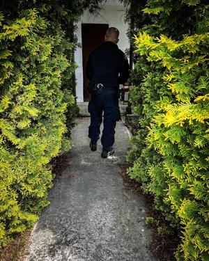 Policjant idzie w kierunku posesji, z dwóch stron otaczają go krzaki.