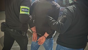 Zdjęcie kolorowe przedstawiające zatrzymanego prowadzonego przez policjantów kryminalnych.