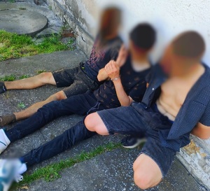 Zdjęcie kolorowe przedstawiające trzech mężczyzn siedzących ma ziemi, zakutych w kajdanki.