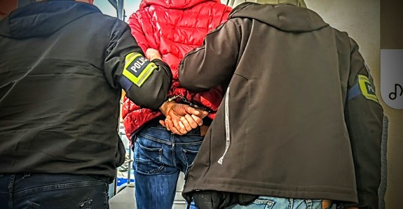 Zdjęcie przedstawia zatrzymanego prowadzonego przez policjantów kryminalnych.