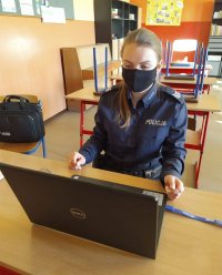 Policjantka siedzi w klasie szkolnej przed biurkiem, na którym stoi laptop