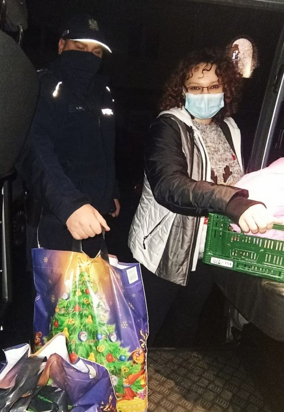 Po lewej stronie stoi umundurowany policjant, w ręku trzyma torbę ze świątecznym motywem. Po prawej stronie stoi kobieta, w rękach trzyma plastikowy kosz wypełniony przedmiotami.