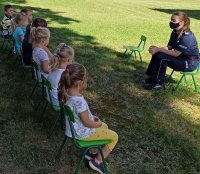 Zdjęcie przedstawia policjantkę, która  przeprowadza zajęcia dydaktyczne z przedszkolakami. Zajęcia odbywają się w okresie letnim, na świeżym powietrzu. Uczestnicy zajęć siedzą na krzesełkach z zachowaniem bezpiecznej odległości.