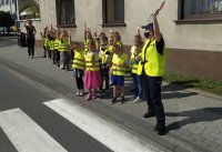 Zdjęcie przedstawia grupe przedszkolaków wraz z policjantka oraz nauczycielka. Uczestnicy zajęć znajdują się na drodze, w rejonie przejścia dla pieszych. Ubrani są w kamizelki odblaskowe, ich ręce są podniesione. Policjantka trzyma w reku tarczę do zatrzymywania pojazdów.