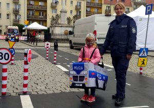 zdjęcie małej dziewczynki i policjantki. dziewczynka ma na sobie założony samochodzik policyjny