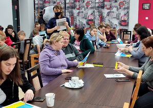 dzieci i ich mamy siedzą w sali, policjantka i sierż. Prudek rozdają elementy odblaskowe, kolorowanki i polsko-ukraińskie materiały informacyjne