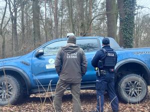 Grafika przedstawia pojazdy policji i straży leśnej. W tle znajduje się las, na terenie którego pełniona jest służba przez funkcjonariuszy obydwu służb.