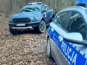 Grafika przedstawia pojazdy policji i straży leśnej. W tle znajduje się las, na terenie którego pełniona jest służba przez funkcjonariuszy obydwu służb.