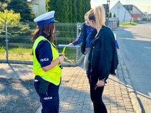 Obraz przedstawia funkcjonariusza wręczającego odblask dzieciom, które zostały przywiezione przez swoich opiekunów do przedszkola