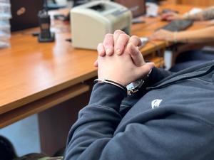 Zdjęcie przedstawia ręce zatrzymanego mężczyzny. Na rękach założone są kajdanki. W tle znajduje się biurko.