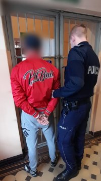 policjant,  zatrzymany mężczyzna w czerwonej kurtce i szarych spodniach, drzwi w komendzie szklane z kratą