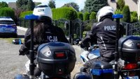 policjant i policjantka na motocyklu służbowym, w tle  budynek jednorodzinny, krzewy i radiowóz