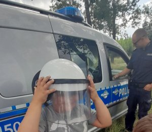 dziecko w hełmie policyjnym, w tle radiowóz i policjant