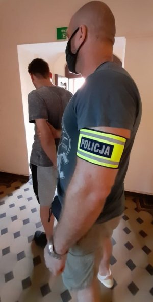 nieumundurowany policjant z zatrzymanym, zatrzymany mężczyzna ma założone kajdanki na ręce trzymane do tyłu, w tle hol Komendy Powiatowej Policji w Krapkowicach