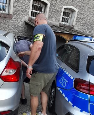 policjant nieumundurowany wprowadza do radiowozu  nieoznakowanego zatrzymanego mężczyznę, samochód koloru srebrnego, obok radiowóz oznakowany