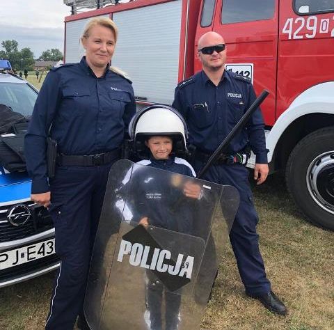 Policjantka i policjant stoją przed radiowozem z chłopcem ubranym w policyjny sprzęt