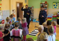 Policjant trzyma w ręce czerwony, żółty i zielony puzzel, przy pomocy których tłumaczy dzieciom zasady poruszania się gdy jest sygnalizacja świetlna.