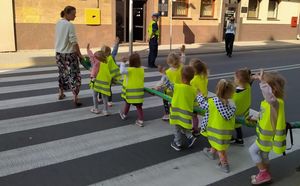 Dzieci ubrane w żółte kamizelki odblaskowe przechodzą przez jezdnię po pasach. Przed nimi policjantka ruchu drogowego.