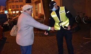 Policjant w kamizelce odblaskowej zakłada odblaskową opaskę kobiecie na prawe przedramię.